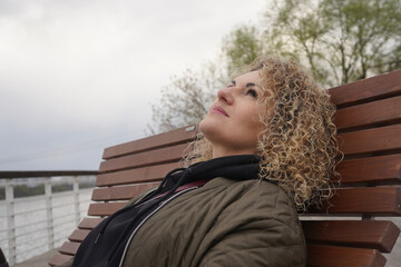 Natural curly blond hair young woman resting on the bench in the park near the river in cold weather