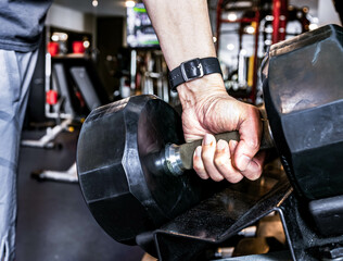 Closeup hand holding black iron dumbbell, muscular build activity. Man taking weights from rack in gym, selective focus. Indoor fitness with blurred workout equipment background. Strong and healthy.