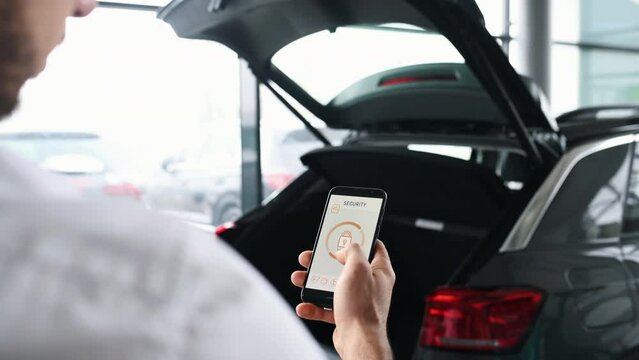 Rear view of man that closing car's trunk by smartphone