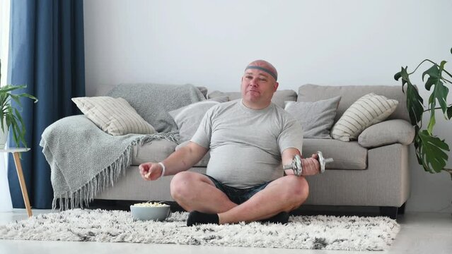 Eating popcorn and lifting weight. Man with overweight is indoors at home