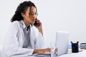 African american mid adult woman talking over smart phone and using laptop on desk, copy space