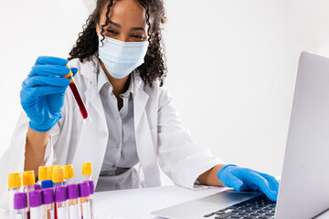 African american mid adult woman wearing mask holding test tube and using laptop on white background