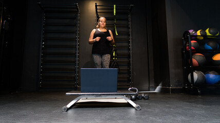 mujer joven de pie en gimnasio frente a un portátil hace ejercicio y toma clases online