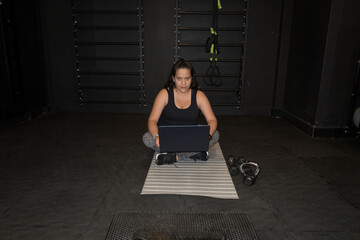mujer joven sentada en una colchoneta de gimnasia con un portátil en las piernas mira a la cámara...
