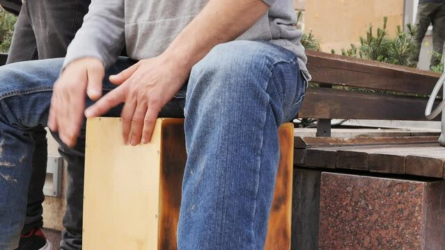 A street drummer taps the cajon with his palms while sitting on it. Playing the cajon with the palms of the tapas. Slow motion, street art