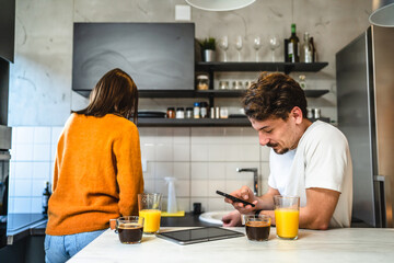Two people young couple adult man and woman husband and wife or boyfriend with girlfriend in the kitchen in the morning with coffee juice mobile phone and digital tablet talking and chat daily routine