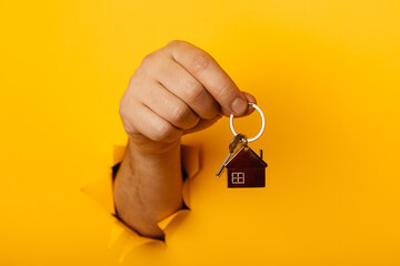 Male hand with metal house and key through a hole in yellow paper wall. House sale and rent concept