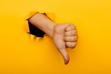 Hand showing a thumb down through ripped hole in yellow paper background. Concept of dislike and disapproval gesture