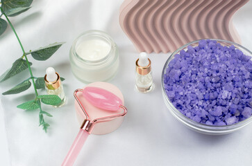 Rose quartz facial roller with face oil, face moisturizer and lavender sea salt on a white table background. Facial massage kit for lifting massage therapy, home spa, mock up