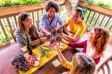 happy multiratial group of friends making a celebratory toast with wine on a picnic table outdoor in the terrace. people gathering in a summer happy hour laughing in a party. joy and lifestyle concept