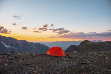 Scenic alpine landscape with tent at very high altitude with view to large mountains in orange dawn sky. Vivid orange tent with awesome view to high mountain range under cloudy sky in sunset colors.