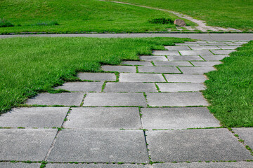 park path made of square tiles overgrown with grass in a parkland with a green lawn close up of the way on park with meadow and asphalt road lit by sun, nobody.