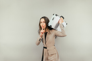 Beautiful business woman in a suit with a metal case of money standing on a beige background shows a gesture of silence to the camera.