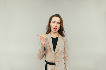 Surprised lady in a beige suit stands on a beige background, looks into the camera with a dissatisfied face and points a finger to the side.