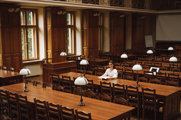 Attractive girl student in a white blouse sitting alone in an atmospheric public library with a book and looking at the camera