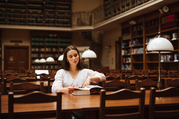 Stylish woman in a white blouse sitting at a long table in an old public library and reading a book with a serious face