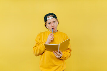 Pensive guy student in headphones with a notebook isolated on a yellow background, looking at the camera with a serious face and biting a pen.