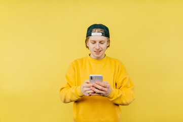 Positive man in a cap and casual clothes stands on a yellow background with a smile on his face and uses a smartphone.