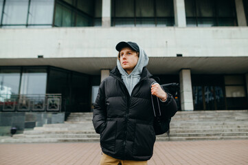 Portrait of a stylish young man in warm clothes walking on the street with a backpack on his back, looking away with a serious face on the background of the building