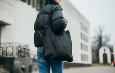 Close photo of a woman in a warm black jacket and with an eco bag in her hands walking down the...