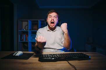Joyful young man sitting at night at the computer at work and emotionally happy with a smile on his...