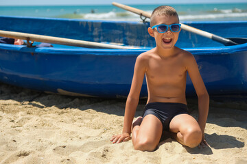 A young boy in a blue boat on the ocean. A child in sunglasses on the beach near the shore. © alexeytsurkan