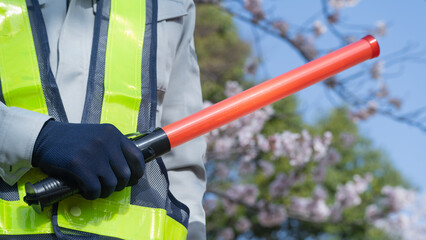 桜の背景で作業服で誘導棒を持つ人｜建設業・警備業