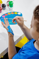A little preschooler sits at a table, holding a brush in his hand and painting paints his palm. Preschooler development.