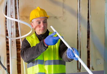 Caucasian electrician in vest and helmet standing with cable duct in hands and looking through it.