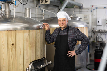 Confident successful brewer in black apron standing in craft brewery against background of fermentation tanks..