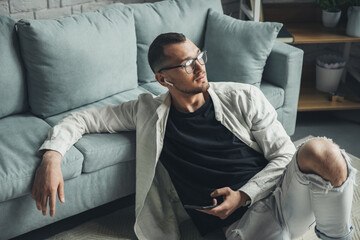Fototapeta premium Man in wireless earphones sitting on the floor near the sofa enjoy good music, play tracks on cell. Flat portrait. Mobile communication.