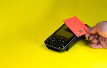 A hand holds a contactless bank card near at the terminal in a yellow background to make a purchase...