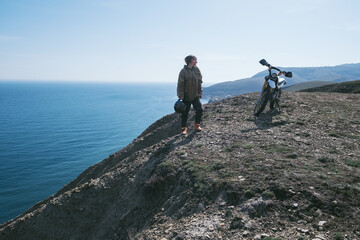 Girl motorcyclist standing on the edge of  cliff holding helmet in her hand, resting after...