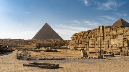 Archaeological complex on the Giza plateau. Dilapidated stone walls are visible. Horses are standing on the tracks. The Great Pyramid of Cheops rises against a background of blue sky and clouds. Egypt - obrazy, fototapety, plakaty