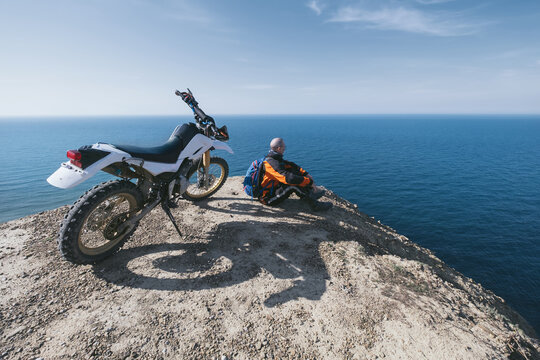  male motorcyclist with grey hair sitting near his dirt motorcycle on steep mountain, resting after difficult drivivng