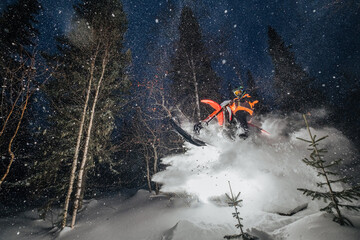 Motorcyclist jumping on snowbike in snow splashes in winter forest, night portrait by flash light
