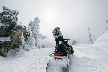 Female riding snowmobile in fabulous winter forest, rocks and pine trees covered with snow, winter haze on top of the mountains