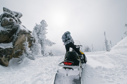Female riding snowmobile in fabulous winter forest, rocks and pine trees covered with snow, winter haze on top of the mountains
