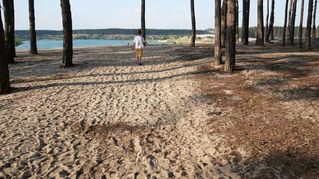 A 5-6 year old boy walks along a sandy path in a pine forest. In front of him is a cliff and a lake with blue water.