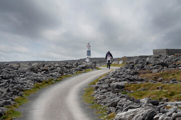 Family with children cycling on small narrow country road in rough stone terrain to a lighthouse. cloudy sky. Inisheer, Aran island, county Galway, Ireland. Irish landscape. Adventure concept.