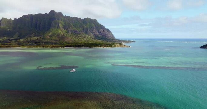 Flying towards Koolau Mountains and white boat on calm colorful ocean with coral reefs in Kaneohe Bay, Oahu Island, Hawaii