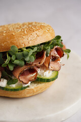 A bagel sandwich with pastrami, cucumber slices, watercress salad and ricotta on round marble board