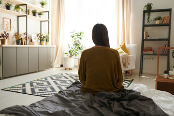 Rear view of lonely woman in cardigan sitting on bed in cozy flat and missing life before...