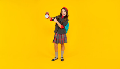 amazed teen girl with school bag pointing finger on alarm clock on yellow background, time.