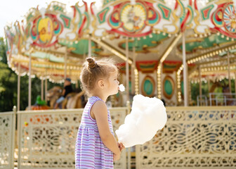 Fototapeta premium Funny blonde girl in colorful dress eating cotton candy at the carnival