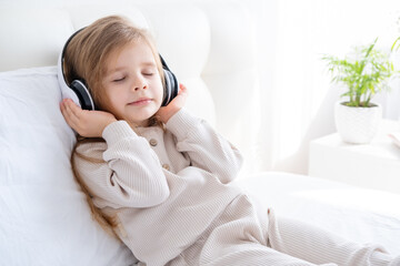 happy little kid caucasian girl with closed eyes in pyjama in wireless headphones on bed