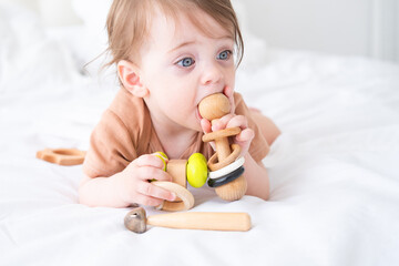 baby girl with blue eyes in brown bodysuit playing with wooden toys on white bedding on bed