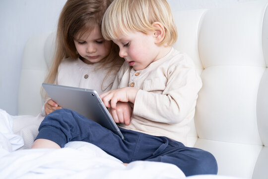two cute kids girl and boy brother and sister using tablet sitting on bed at light modern bedroom