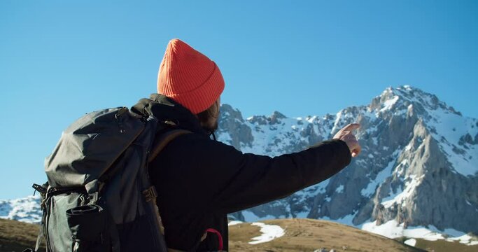 Portrait of excited young traveller man with backpack on his mountain adventure. Hiker showing with his hand the way to mountain peaks. Person pointing with arm epic views. 