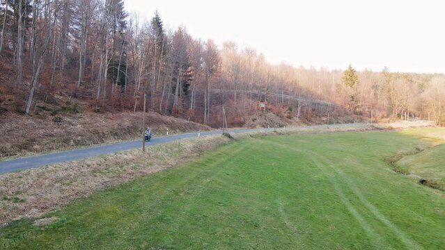 Motorcyclist driving along a narrow, empty forest road next to a green meadow. Static wide angle view